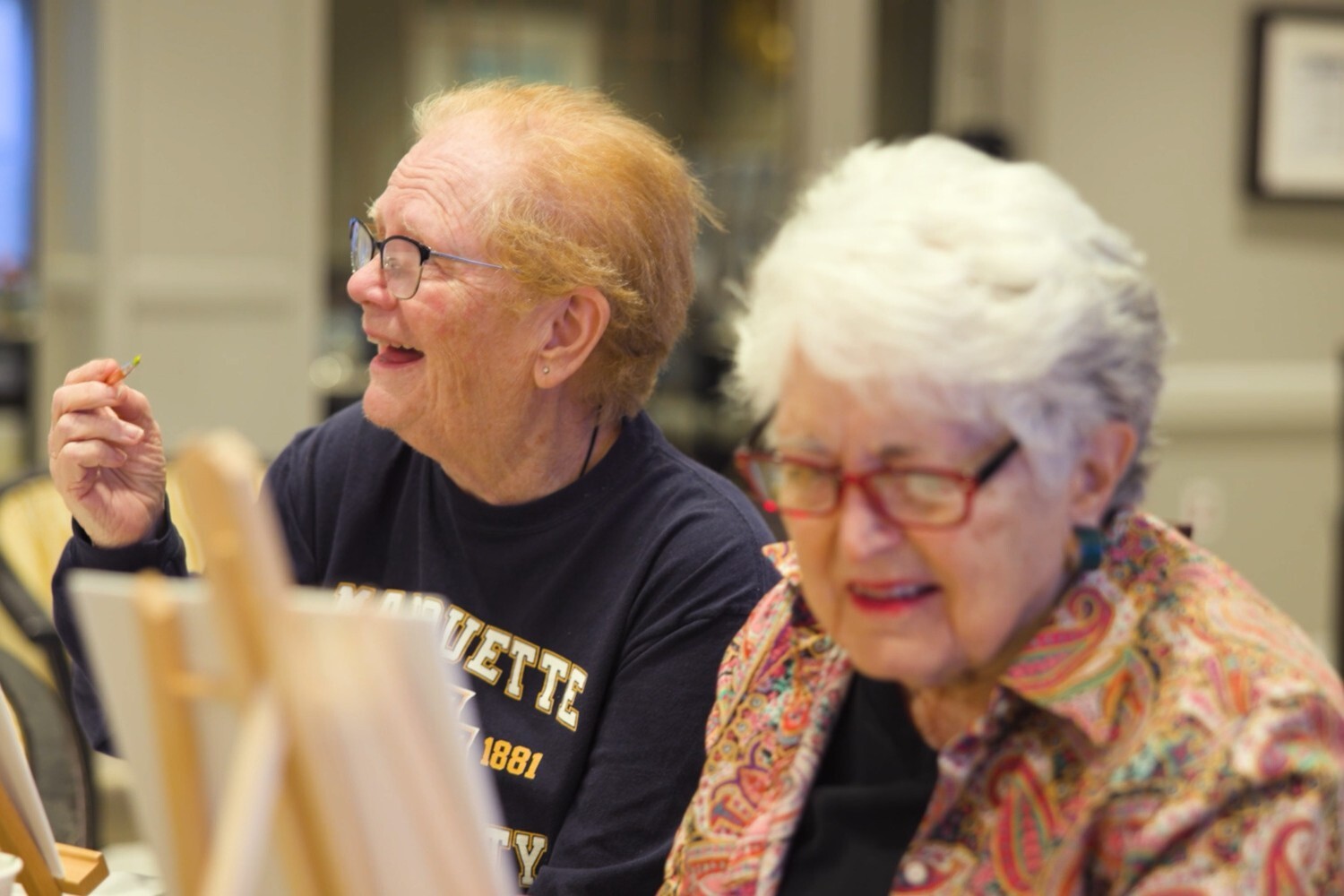 Two residents at Arbor Terrace Glenview senior living smile while participating in a creative painting class, embracing the active senior lifestyle Glenview offers.